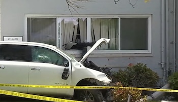 Camión pickup blanco severamente dañado con el capó abierto, empotrado contra la fachada de un edificio con una ventana rota. Cinta policial amarilla visible