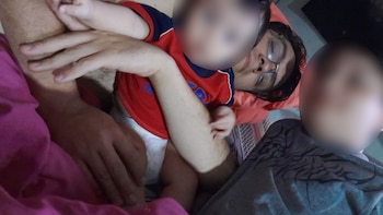 Primer plano de una persona con gafas acostada con los ojos cerrados, sosteniendo a un bebé con pañal y camiseta roja. Se ven sus manos y las del bebé