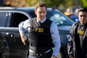 Dos agentes del FBI, uno con chaleco antibalas y pistola enfundada, y otro con chaqueta negra, frente a un SUV oscuro con luces de emergencia