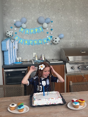 Niña con trenzas y flor en el cabello frente a una torta de cumpleaños, con globos, piñata de camiseta argentina y adornos de fútbol al fondo