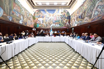 Personas sentadas alrededor de mesas con manteles blancos en un salón con micrófonos, pantallas y murales en las paredes