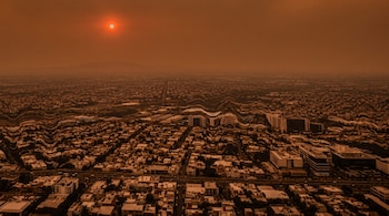 Vista aérea de una ciudad densamente poblada bajo un cielo naranja y brumoso con un sol brillante, mostrando edificios y calles con un efecto ondulado.
