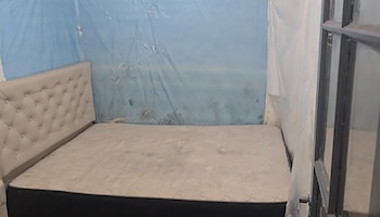 Cama con cabecero tapizado beige y colchón sobre una base oscura en una habitación. Las paredes están cubiertas con plástico protector azul y transparente, y hay una ventana a la derecha