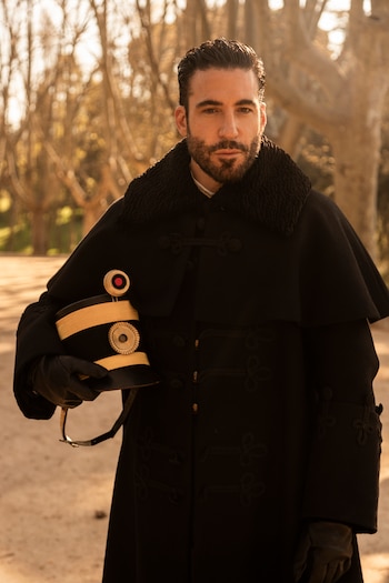 Hombre con barba, abrigo negro, guantes negros y sombrero militar con detalles dorados, de pie en un exterior con árboles