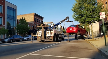 Una grúa blanca y azul levanta un auto rojo en una calle urbana. Se ven edificios, árboles y un letrero de 'No Estacionar'.