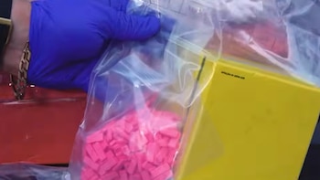 Una mano con guante de látex azul sostiene una bolsa de plástico transparente con pastillas de color rosado junto a una caja amarilla