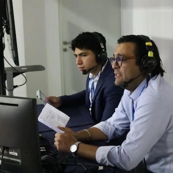 Dos hombres con auriculares y micrófonos sentados frente a monitores; el de la derecha, con gafas y camisa clara, sostiene papeles; el de la izquierda usa traje