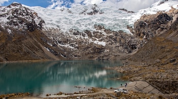 Laguna turquesa frente a un glaciar con rocas, una presa de concreto, un vehículo blanco y varias personas en la orilla.