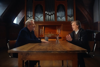 Paul McCartney con camisa blanca y chaqueta oscura, sentado frente a un hombre con gafas y traje oscuro, en una sala con un gran órgano de tubos
