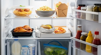 Refrigerador blanco con estantes llenos de sobras: pollo asado, espaguetis con albóndigas, arroz, pizza, patata asada y varios condimentos.