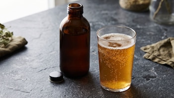 Un vaso con una bebida dorada y espumosa junto a una botella de vidrio ámbar destapada y su tapa sobre una mesa oscura