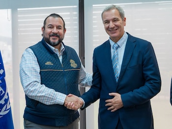 Dos hombres, uno con chaleco oscuro y el otro con traje, se dan la mano. El hombre del chaleco tiene barba y el logo IGSS