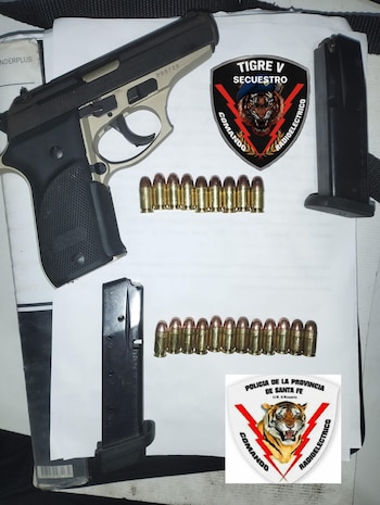 Una pistola bicolor, dos cargadores y diecinueve balas dispuestas en dos filas sobre una superficie blanca, junto a dos insignias policiales