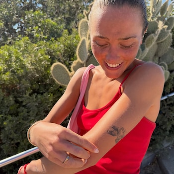 Mujer joven con cabello rubio recogido, top rojo, bolso rosa. Brazos con tatuaje y pulsera. Anillo en dedo anular. Fondo de cactus y vegetación