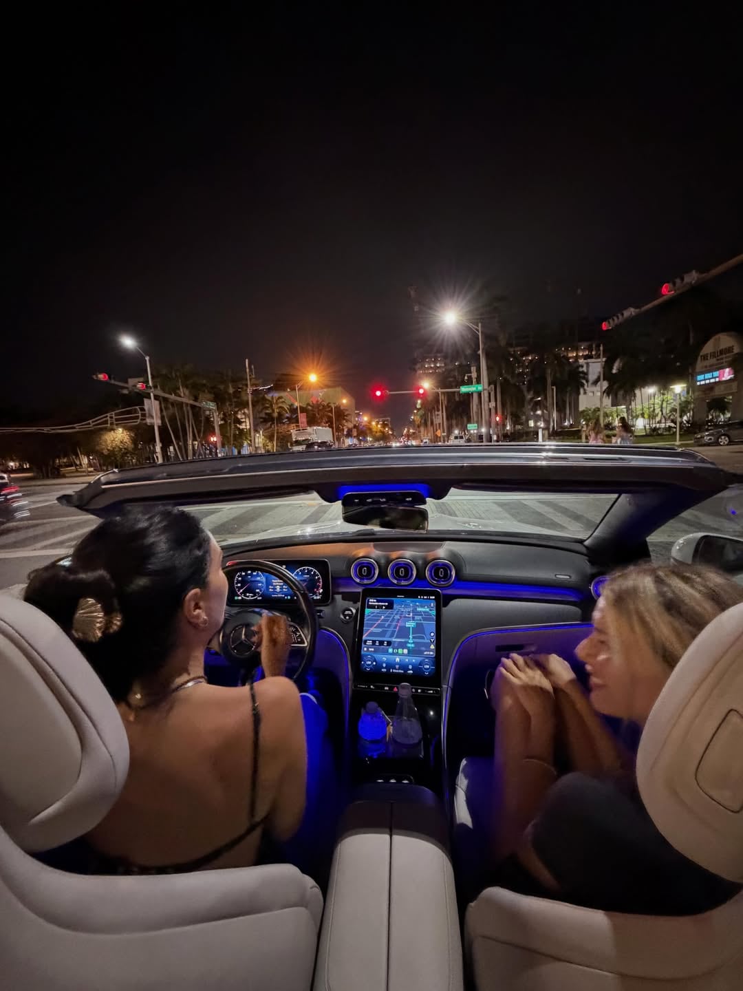 Zaira al volante de un lujoso descapotable por las iluminadas calles de Miami de noche, acompañada de su amiga Guillermina en el asiento del pasajero