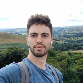 Un hombre joven con barba, camiseta azul y auriculares blancos posa al aire libre ante un paisaje de colinas verdes
