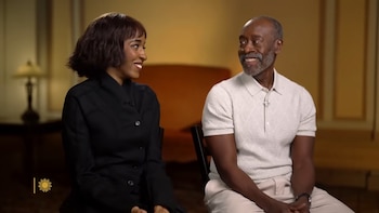 Ayo Edebiri y Don Cheadle sentados, mirándose y sonriendo. Ella lleva una chaqueta negra y él un polo blanco con pantalón claro