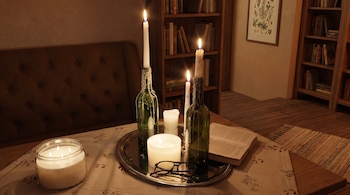 Mesa con velas encendidas en botellas de vidrio, un libro abierto, anteojos y estantes con libros en el fondo.