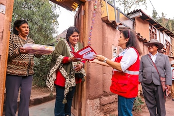 Una mujer con chaleco rojo entrega una carpeta abierta con documentos a otra mujer frente a la puerta de madera de una casa de adobe