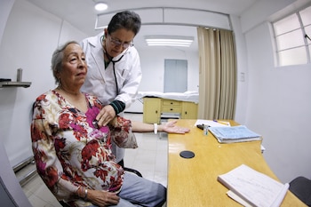 Una doctora con bata blanca y estetoscopio examina el pecho de una paciente mayor sentada en un consultorio médico con un escritorio y una camilla