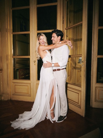 Una mujer rubia y un hombre sonrientes se abrazan frente a puertas de madera ornamentadas y marcos dorados, en un interior elegante