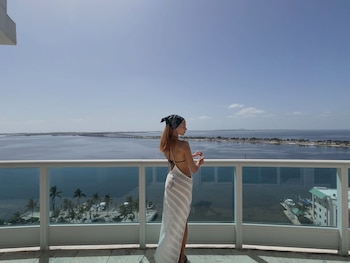 Una mujer de perfil con pañuelo en la cabeza y falda blanca posa junto a la barandilla de cristal de un balcón frente al mar