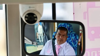 Un espejo retrovisor refleja a un conductor con camisa blanca y corbata rosada junto a una cámara de seguridad en el interior de un bus
