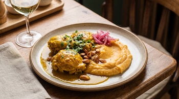Un plato contiene tres porciones de pollo, puré amarillo, cebolla morada encurtida y almendras. Un vaso con vino blanco y una servilleta están al lado.