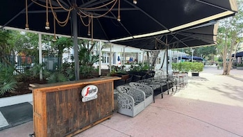 Vista de una terraza de café al aire libre con un bar de madera, sombrillas negras y sillas en una acera, rodeada de vegetación