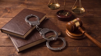 Dos libros cerrados de cubierta marrón, unas esposas de metal, un mazo de madera y una balanza de justicia de latón sobre una superficie de madera.