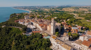 Vista aérea de un pueblo con casas de tejados rojos, una iglesia con torre campanario, un área arbolada y el mar.