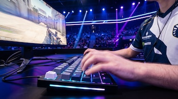Primer plano de las manos de un jugador de eSports sobre un teclado retroiluminado con luces de neón azules y moradas, mientras juega un videojuego en un monitor en un estadio abarrotado.