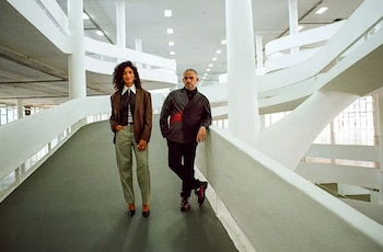 Amanda Carneiro, con cabello rizado y chaqueta marrón, y Raphael Fonseca, con chaqueta oscura, de pie en una rampa blanca dentro de un edificio moderno