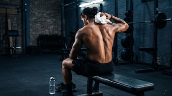 Hombre de espaldas sin camiseta sentado en un banco de gimnasio secándose el cuello con una toalla blanca junto a una botella de agua.