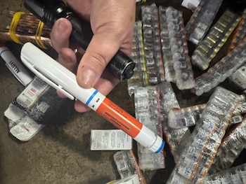 Mano sostiene linterna negra y pluma inyectora blanca y naranja. Alrededor hay numerosos paquetes blíster sellados con viales farmacéuticos