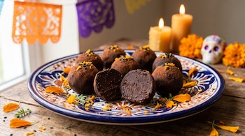 Trufas de chocolate en un plato decorado, con una cortada a la mitad, pétalos naranjas, velas encendidas, papel picado y una calavera al fondo.