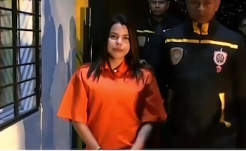 Una mujer joven con cabello oscuro y camisa naranja mira a cámara, mientras es escoltada por dos hombres en uniforme oscuro en un interior