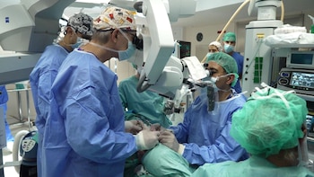 Cirujanos con uniformes quirúrgicos azules y verdes operan en un quirófano moderno, utilizando un microscopio y diversos equipos médicos