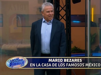 Mario Bezares viste un saco oscuro y camisa azul claro frente a un letrero del programa La Casa de los Famosos México