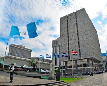 Un edificio de hormigón gris con textura. La bandera de Guatemala ondea prominentemente. Varias otras banderas y un oficial de uniforme de pie en la plaza