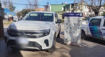 Una camioneta blanca estacionada junto a un cartel institucional de la policía y una patrulla en una calle residencial