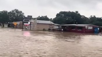 Agua de inundación marrón cubre una calle frente a varias edificaciones y árboles bajo un cielo nublado