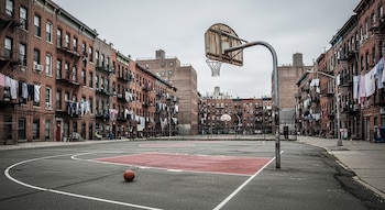 Una cancha de baloncesto al aire libre con un balón, rodeada por edificios de ladrillo con escaleras de incendio y ropa tendida bajo un cielo nublado.