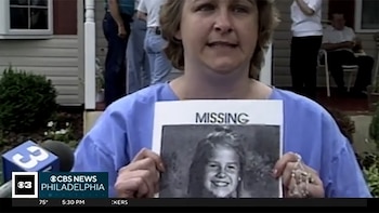 Mujer de mediana edad con camisa azul sostiene cartel de "MISSING" con foto en blanco y negro de una niña sonriente. Al fondo, micrófono y parte de una casa