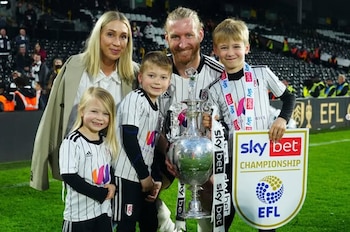 Tim Ream con barba, su esposa rubia y sus tres hijos pequeños posan en un campo de fútbol con un trofeo plateado y un cartel del Sky Bet Championship