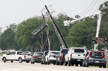 Vista de un poste de electricidad inclinado en una carretera con un trabajador en una grúa aérea reparando cables, y una fila de coches esperando
