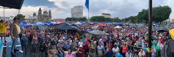 Mickey Singh, en el escenario, levanta el puño hacia una inmensa multitud. El fondo muestra carpas de colores, árboles y edificios de la ciudad bajo un cielo nublado