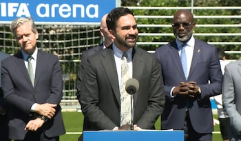 Tres hombres con traje y corbata, incluyendo uno que habla en un micrófono, de pie frente a una portería de fútbol y un cartel azul de FIFA