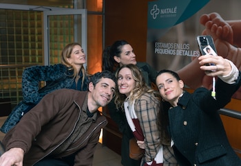Cinco personas sonrientes, tres mujeres y dos hombres, posan para una selfie tomada por una mujer con un teléfono móvil en un interior luminoso