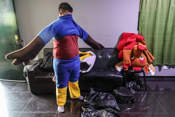 Hombre de espaldas con traje de colores, poniéndose o quitándose guantes grandes de disfraz. Una cabeza de mascota marrón y roja está en una silla
